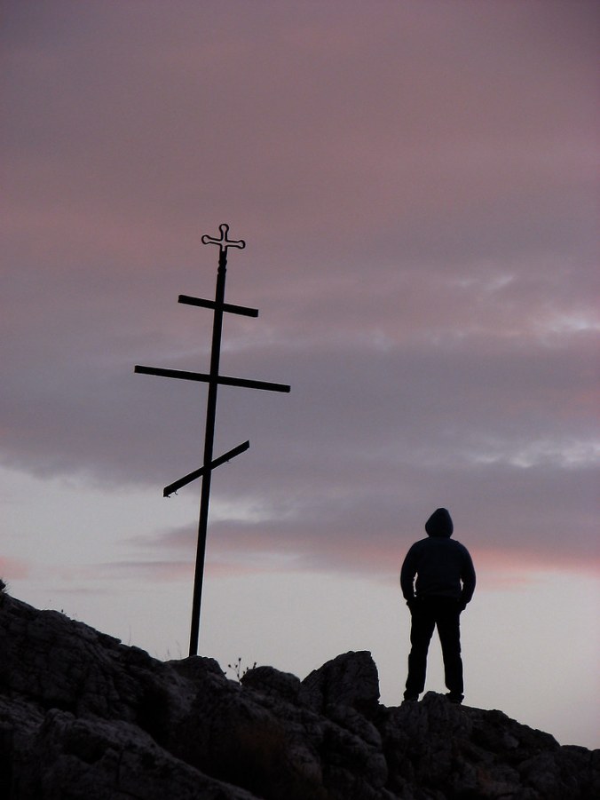 Silhouette of a young man standing next to a cross on sunset